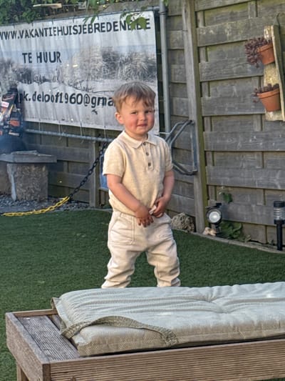 The image, oriented vertically, captures a young child standing in an outdoor setting, likely a backyard or patio. The child, a fair-haired toddler, is dressed in a light-colored collared shirt with buttons and matching long trousers. They are looking towards the viewer with a calm, slightly smiling expression, one hand resting on their stomach.
In the background, a wooden slatted fence serves as a backdrop. Attached to the fence are two small terracotta pots, each containing what appear to be dried or dead plants, and several solar-powered garden lights. A large white banner or sign is also affixed to the fence, positioned behind and to the left of the child. The ground is covered with green artificial turf, giving the area a neat, maintained appearance. To the right of the child, a piece of outdoor furniture, possibly a sofa or bench, with a light-colored, tufted cushion is visible. A metal chain, part black and part yellow, lies on the turf near the child's feet, extending towards the fence.
Visible text on the banner includes "WWW.VAKANTIEHUISJEBREDENE" (likely part of a website address for a vacation home), "TE HUUR" (Dutch for "FOR RENT"), and "nk.deloof1960@gma" (an incomplete email address, likely @gmail.com).
The photograph appears to be taken during the daytime under soft, natural lighting, suggesting either an overcast day or the softer light of morning or late afternoon. The atmosphere is relaxed and candid. The location is in Bredene, Belgium, as indicated by the context and the banner text.