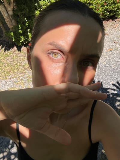 This media file is a close-up, low-angle, outdoor photograph of a person's face, primarily focused on their eyes and hand. The person appears to be an adult with light skin and dark hair. Their face is partially obscured by their right hand, which is held up to their face, with the fingers splayed and partially covering the mouth and nose area. The person's left eye is clearly visible, showing a hazel iris with some yellow and green tones. The right eye is partially visible, also appearing to be hazel. Sunlight casts a diagonal shadow across the left side of the person's face and forehead, creating a dramatic contrast with the illuminated right side.
The setting appears to be outdoors, with elements of nature visible in the background, including green foliage, what looks like gravel or small stones on the ground, and tree trunks. The lighting suggests it is daytime, likely a sunny day, given the strong shadows. The overall mood conveyed is somewhat mysterious or introspective, possibly due to the partial concealment of the face and the intense gaze of the visible eye. There is no visible text in the image. The location context provided is Baan Mae Nam, Thailand.