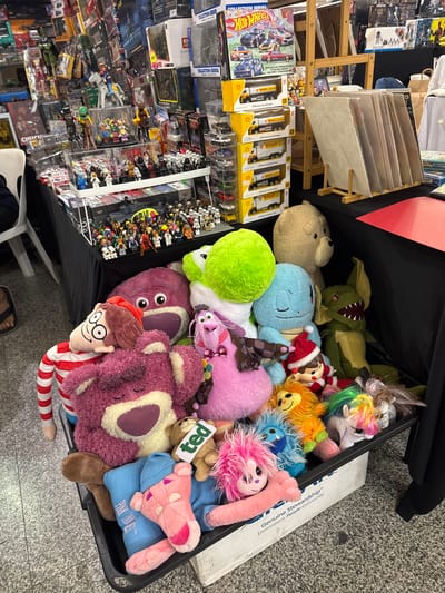 The image shows a table laden with a variety of collectible toys and merchandise, likely at a bazaar or convention in Kuala Lumpur, Malaysia. Dominating the foreground is a large black plastic bin filled with plush toys, including a large purple bear, a pink bear, a blue character resembling Squirtle, a green creature with sharp teeth, and a white unicorn with a rainbow mane. A plush toy of Waldo, a character from the "Where's Waldo?" books, is visible on the left, wearing his signature red and white striped shirt. Behind the bin, on the table, are more toys and merchandise, including a display of action figures and smaller collectible items. Further back, shelves are stocked with boxed toys and merchandise, featuring brands like Hot Wheels and various comic book or movie-related figures. The overall environment appears to be an indoor market or exhibition hall with a tiled floor.
