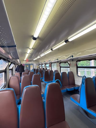 The image displays the interior of a commuter train car, likely in Komarovo, Russia, rotated 90 degrees clockwise. The scene captures a long aisle with rows of seats predominantly empty. The seats feature distinct brown upholstered cushions and bright blue metal frames or bases, arranged in pairs facing each other in some sections and forward-facing in others.
On the left side of the frame, serving as the "ceiling" in a normal orientation, are multiple long, bright fluorescent or LED tube lights that illuminate the car. These lights are mounted on a cream-colored panel system, which also features sections with small, uniform perforations, likely for ventilation or speakers.
The "floor" of the train car, seen on the right side of the image, has a light grey finish. Windows line the right side of the car, offering glimpses of green foliage and a metallic fence or railing outside, suggesting the train is either stopped at a station or moving through a natural landscape. Small red "No Smoking" symbols are visible on stickers near the windows.
Towards the far end of the car, a person dressed in dark clothing is partially visible, either sitting or standing. Above the far door, a digital display shows red numbers, possibly "23:13" and "09", which could indicate time or other train information. The overall impression is one of a clean, well-lit, and functional public transport space during what appears to be daytime, given the natural light from the windows.