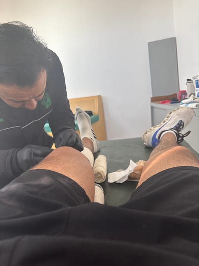 A person with black gloves is wrapping bandages on their leg, which is resting on a green surface. Beside the wrapped area are two rolls of bandages. Above, another leg wearing a sock and shoe sits on the same surface. The person wrapping the bandage is wearing a black and green athletic shirt. Tissue paper is scattered on the surface near the wrapped legs and bandages. A shelf with various items is visible in the background.