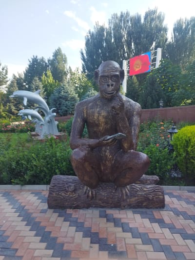 The image shows a statue of a monkey sitting on a log, holding a smartphone. The statue is in a park-like setting with lush green bushes and trees in the background. There is also a statue of dolphins jumping out of water to the left of the monkey statue. A flag with a red field and a yellow sun symbol is visible in the background. The ground in front of the statue is paved with colorful interlocking bricks in a diagonal pattern. The scene appears to be in a public space, likely a park or plaza in Kenbulun, Kyrgyzstan, given the presence of the national flag. The lighting suggests it is daytime, with sunlight illuminating the scene.