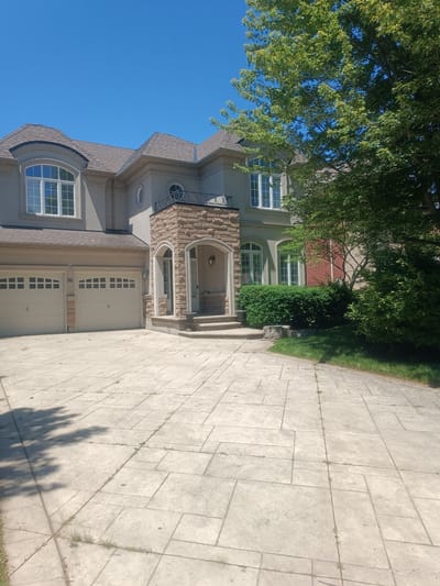 A wide shot captures a light grey stucco, two-story luxury home located at 96 Oak Ridge Trail in Oakville, Ontario, Canada. The house features a multi-pitched asphalt shingle roof, several arched windows with white trim, and a prominent stone-clad entrance with an arched opening. Two beige garage doors are visible on the left side of the house. A large, leafy green tree with dappled shade casts shadows across the pale grey concrete driveway, which is stamped with a large tile pattern. The driveway has some sparse weeds growing in the cracks. The sky is a clear, bright blue. The overall scene depicts a well-maintained, upscale residential property on a sunny day.