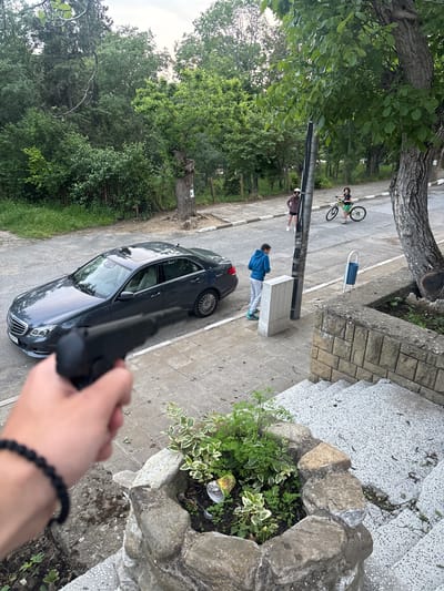 The image, taken from an elevated position, shows a street scene with a parked dark-colored car. A person in a blue jacket and grey pants stands next to a light-colored structure on the sidewalk. Further down the street, another individual is walking while a third person is riding a bicycle. A hand holding a dark object, possibly a gun, is visible in the foreground, obscuring part of the car. The setting appears to be in a residential area with trees and greenery visible. Based on the prompt, the location is in Goritsa, Bulgaria.