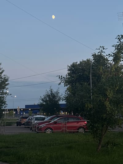 The image depicts a street scene at dusk in Magnitogorsk, Russia. The sky is a pale blue with some light cloud cover, and a visible moon is present, indicating it is likely evening. A few power lines crisscross the sky. On the right side of the image, a row of parked cars is visible, including a red car prominently in the foreground. The cars are parked along a grassy area with a metal railing separating the grass from what appears to be a sidewalk or parking area. Trees with green foliage are scattered throughout the scene, some casting shadows. In the background, a blue building with signage is partially visible, along with streetlights. The overall impression is a quiet evening in a residential or commercial area. There are no people visible in the image.
