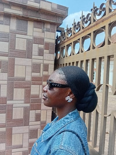 An individual, presumably female, with dark skin, is depicted in profile facing left in Ejule, Nigeria. She wears a black fabric head wrap styled into a bun at the back, black framed sunglasses, and a light blue denim jacket. A silver-colored, bow-shaped earring is visible on her right ear, and a dark strap crosses her right shoulder. To her left is a wall constructed with geometrically patterned tiles in shades of brown and beige, topped with a decorative cornice. To her right is a tall, brown metal gate featuring intricate scrollwork, finials, and a section with circular cutouts. Through the gate, a glimpse of a light-colored building with a reddish roof is visible under a partly cloudy blue sky.