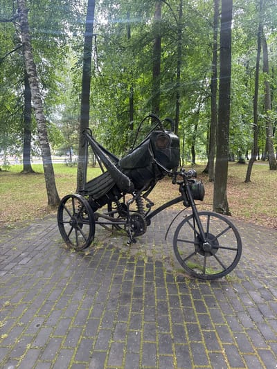 This media file shows a sculpture of a diver's helmet and suit mounted on a three-wheeled vehicle, possibly a custom-made bicycle or a small, wheeled platform. The sculpture is made of dark metal and features intricate mechanical details, including springs and articulated parts. The diver's helmet has a distinct, round faceplate with mesh detailing. The vehicle has two large spoked wheels at the rear and a smaller wheel at the front.
The sculpture is situated outdoors on a paved walkway made of dark rectangular bricks with moss growing in the gaps between them. The setting is a park or wooded area, with numerous trees visible in the background, their leaves creating a lush green canopy. Sunlight filters through the leaves, casting dappled light. The grass in the background is green and appears well-maintained. The overall scene suggests a public space, possibly an art installation in a park.
There is no visible text in the image, and the time of day appears to be daytime, likely morning or afternoon, given the lighting conditions. The weather seems pleasant, with clear skies and sunlight. The image evokes a sense of artistry and perhaps a touch of whimsy, given the unusual combination of a diver's suit and a wheeled vehicle. The location context provided is Sarov, Russia, suggesting this is an outdoor exhibit in that city.