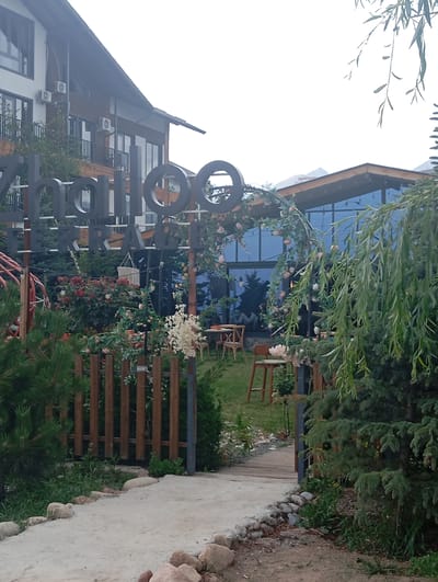 The image shows the exterior of a building in Бактуу-Долоноту, Kyrgyzstan. The building has a modern architectural style with multiple stories, large windows, and balconies. In the foreground, there is a pathway leading to the building, bordered by rocks and greenery. A wooden fence separates the pathway from a garden area with tables and chairs set up for outdoor dining. A large sign with stylized letters is visible in front of the building. The environment appears to be a resort or restaurant setting, possibly near a natural landscape given the presence of trees and the faint outline of mountains in the background. The overall impression is a serene and inviting outdoor space.
