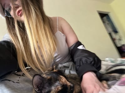 A woman with blonde-streaked brown hair is holding a tortoiseshell dog. The woman is wearing glasses, a black jacket with white trim, a gray sweatshirt, and a white tank top. The background is a light yellow wall, and a dark object is partially visible on the left side of the frame. There is no indication that the image was taken in Pushchino, Russia.