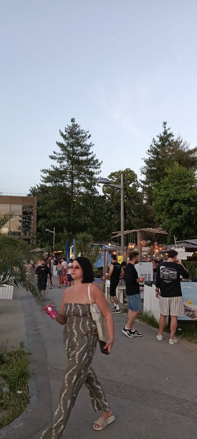 The image depicts a scene on a warm evening at the "Kస్తున్నాయి" beach bar, located on the Donauufer (Danube riverbank) in Linz, Austria. A woman in the foreground is walking away from the camera, holding a pink can and wearing a green and white striped one-piece outfit and beige slides. She carries a cream-colored bag over her shoulder. Behind her, several people are present. To the left, a palm tree and white fencing are visible. The background features a building with large windows, trees, and the beach bar area with wooden structures, string lights, and people socializing. The sky is clear and light blue.