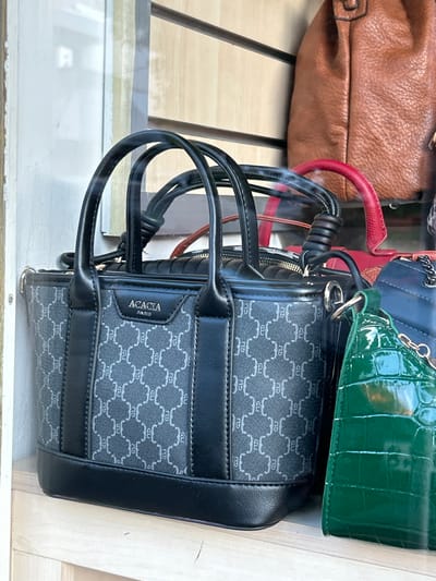 The image captures a display of handbags, likely within a retail setting in Koridallos, Greece. The photo is rotated 90 degrees clockwise, presenting the items horizontally.
The primary subject is a dark grey and black small handbag with a distinctive repeating monogram-like pattern, possibly consisting of stylized 'R' or 'B' letters. The bag features black leather accents, including handles and trim. A rectangular label on the side clearly displays the brand name "ACACIA PARIS". This bag appears to be made of a canvas-like material with structured leather elements.
Behind and partially obscuring the main bag are several other handbags. To the upper left, a brown, soft-textured leather-like handbag is visible. Towards the top right, a vibrant green patent leather-like handbag with a crocodile skin texture is prominent. A small red handle from another bag is also visible. Further back, parts of a dark blue quilted bag can be discerned.
The bags are arranged on what appears to be a wooden slatted surface, possibly a shelf or part of a display case. There is a noticeable glare or reflection on a transparent surface in the foreground, suggesting the image was taken through glass, such as a shop window. The overall scene depicts a retail merchandising display. No people are visible, and the time of day or weather cannot be determined.