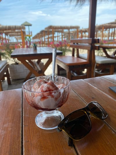 The image captures a moment at a likely seaside establishment in Ulcinj, Montenegro. The scene is dominated by a wooden table on which sits a glass of strawberry ice cream, complete with a spoon, and a pair of Ray-Ban sunglasses. The background reveals a row of tables and chairs under thatched roof structures, indicating an open-air dining area or beachside setting. The sky is visible between the structures, suggesting a sunny day. The scene is quiet, with no people immediately visible. The focus is on the personal items and dessert, perhaps awaiting consumption.