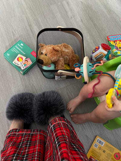 This overhead image captures an intimate indoor scene on a light gray wooden or laminate floor in Dubovka, Russia. The primary subjects are a baby and an adult engaged in a play session.

The baby, visible from the mid-thigh down, is seated in a green plastic support chair or potty. They are holding a yellow rattle in their right hand, while their bare feet rest on the floor. An interactive toy, featuring a light blue and white base with flexible, colorful tubes and rings, is positioned in front of the baby.

To the right of the frame, an adult's legs and feet are visible, dressed in red and black plaid pajamas with white piping. The adult is wearing dark grey, fluffy slippers.

Various toys and children's items are scattered on the floor. A black wire mesh basket holds a brown stuffed teddy bear and other objects. Next to the basket is a teal-colored box with Russian text, which reads "Умница Бормоталки" (Smarty Babbles) and "Для запуска и развития речи" (For starting and developing speech). The box also shows example sounds "му-му-му" and "бах-бах". Another children's book, partially visible at the top left, has the Russian word "Милашки" (Cuties) on its cover. A yellow book or box at the top right displays a barcode "9785506 09872" and the "EAC O+" mark, indicating its suitability for infants. Another toy, resembling a car dashboard with a red steering wheel and colorful buttons, is also on the floor. The overall scene suggests a comfortable and playful environment.