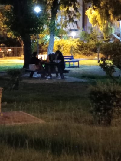In an outdoor urban setting at night, likely a park or public square in Antalya, Turkey, a group of individuals is seated around a picnic table. The scene is dimly lit by overhead streetlights and ambient building lights. The individuals are mostly silhouetted against the background. One person appears to be holding a phone and looking down at it, while others are facing each other, possibly engaged in conversation. A blue park bench is visible in the background. The ground is covered in grass, and trees with foliage are scattered throughout the area. A fence or railing is present on the left side of the image.