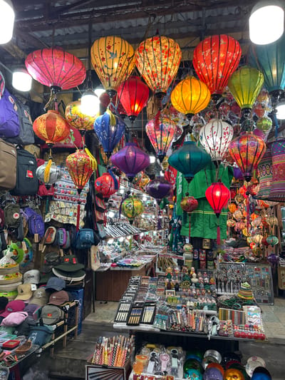The image depicts a vibrant street market scene, likely in Hội An, Vietnam, characterized by an abundance of colorful silk lanterns hanging overhead and displayed on stalls. Numerous stalls are visible, laden with various goods. One stall, prominently featured in the foreground, displays an array of hats of different styles and colors, including baseball caps and conical hats. Adjacent to this, another stall showcases an assortment of items, including chopsticks, carved figurines, beaded necklaces, and other trinkets, suggesting a marketplace offering souvenirs and local crafts. The stalls are constructed with simple materials, some with wooden or metal framing, and are set up along a walkway or street. The lighting appears to be a mix of natural light filtering from above and artificial light from the lanterns and stall lighting, creating a warm and inviting atmosphere. The overall impression is one of a bustling, traditional market, rich in visual detail and local color.