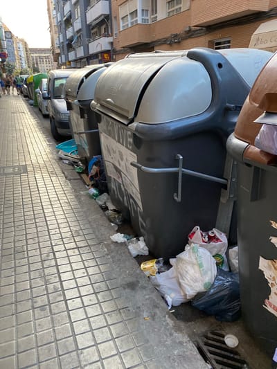 The image depicts a street scene in Valencia, Spain, characterized by overflowing garbage bins and scattered refuse along a tiled sidewalk. In the foreground, a row of large, dark-colored, wheeled garbage bins is prominently displayed, with their lids partially open or closed. The bins are filled with a mix of garbage bags, loose trash, and recyclable materials, including plastic bottles and aluminum cans, some of which are spilling onto the pavement. A white plastic lid rests near a drainage grate.
Adjacent to the bins, a light-colored tiled sidewalk stretches into the background, with a few people walking along it. Further down the street, a line of parked cars is visible, including a silver sedan and several other vehicles. The buildings lining the street are multi-story structures with balconies and windows, suggesting an urban residential or commercial area. The sunlight indicates it is daytime. The overall impression is one of urban neglect, with a significant accumulation of litter detracting from the street's cleanliness.