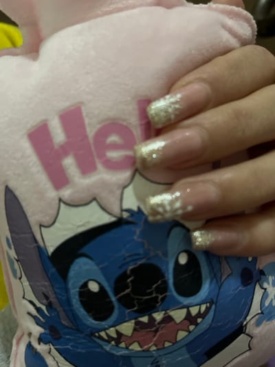 This image, located in Jaramana, Syria, depicts a close-up of a human hand resting upon a plush object. The hand, identified as a right hand, exhibits manicured fingernails with extended, square-tipped acrylic or gel enhancements. These nails feature a clear base, gold glitter applied to the tips in a French manicure style, and a distinct white snowflake or starburst design overlaying the glitter on at least two visible nails.

The plush object beneath the hand is light pink in color and appears to be a pillow or soft toy. It features a printed graphic of the blue animated character "Stitch" from Disney's Lilo & Stitch. Stitch is depicted with large black eyes, a wide open mouth showcasing white teeth, and appears somewhat distressed due to significant cracking and wear on the printed surface. Above the character, the partial word "He," likely part of "Hello," is visible in large pink block letters on the pink fabric, along with a portion of a white speech bubble graphic. The fingers of the hand are in light contact with the plush surface. The background is out of focus, suggesting an indoor, personal environment.