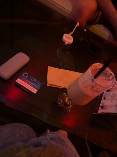 A low-angle, close-up shot captures a dark, reflective surface, likely a table in a dimly lit establishment, with splashes of red and blue light illuminating the scene. On the left, a light-colored, rectangular case rests, positioned above a blue and teal package of TEREA tobacco sticks for IQOS ILUMA, featuring Bulgarian text. To the right of the tobacco packaging, an orange paper napkin is folded. Further to the right, a tall glass filled with a light-colored beverage, possibly a milkshake or a cocktail, contains a dark straw and is tilted to the side. The glass sits on a glossy menu with a Coca-Cola logo visible. In the background, a person's legs and feet are visible, suggesting they are seated. The overall impression is of a relaxed setting, possibly a bar or cafe, in Haskovo, Bulgaria, where the subjects are enjoying drinks and possibly smoking alternatives.