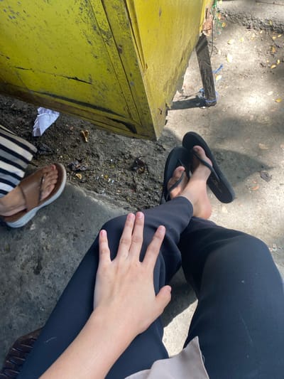 An overhead shot shows two pairs of feet. One person wears black flip-flops and black pants. The other person wears brown sandals and striped pants. The individuals are sitting on a concrete surface with a bright yellow, worn structure behind them. The ground is dusty and has scattered leaves and a crumpled piece of white paper. Sunlight casts shadows. The context of Ta', Indonesia is not ascertainable from the visual information provided.