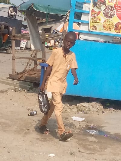 This image depicts a young male subject walking on an unpaved, dusty street in Badore, Nigeria. The individual, appearing to be a boy, wears a light tan, short-sleeved tunic and matching trousers, along with dark sandals. He carries a black plastic bag in his left hand and looks directly towards the camera. The ground shows signs of debris, including a dark brown plastic bottle lying horizontally and a white piece of litter. To the right, a bright blue kiosk or stall displays a multi-panel food advertisement poster featuring items like "Served HOT" and various pasta dishes. To the left, a wooden display frame stands under a large green umbrella, with a light-colored vehicle partially visible in the distant background. The scene is illuminated by direct daylight.