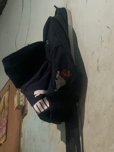 A dark blue Puma backpack is hanging on a wall in what appears to be an indoor setting. The backpack is partially open, revealing some of its contents, including what looks like a small red object. The wall is a textured, light-colored surface with some dark marks and stains. To the left of the backpack, a framed picture or artwork is visible on a wooden surface, and a white electrical box with three circular openings is mounted on the wall. The lighting suggests it might be nighttime or a poorly lit room. The scene is static, with no visible activity. The image quality is slightly grainy. This scene is located in the city of Tacarigua, Venezuela.