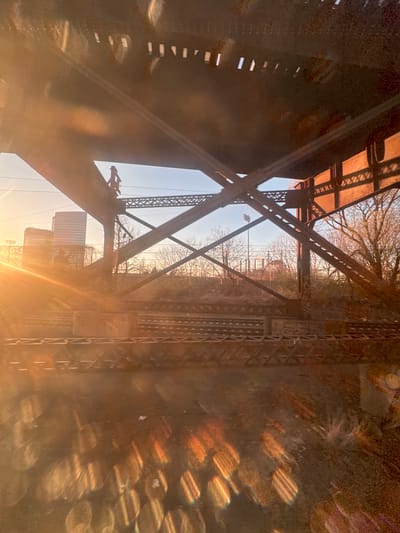 The image depicts an elevated railway bridge structure in Philadelphia, Pennsylvania, likely near the Schuylkill River and 30th Street Station, with the University City skyline visible in the distance. The scene is captured at sunrise or sunset, characterized by a low, warm sun casting significant lens flare from the left.

The foreground and midground are dominated by the rusted, dark brown metal framework of a large bridge, featuring heavy I-beams, diagonal trusses, and riveted plates. Multiple sets of parallel railway tracks run beneath this structure. On a diagonal metal support beam in the upper-left quadrant of the bridge, a silhouetted human figure is visible, appearing to be an adult male wearing a hat or hood and a backpack, positioned as if climbing or traversing the structure.

In the background to the left, several tall, modern urban buildings, including a prominent cylindrical or rounded skyscraper, are silhouetted against the bright horizon, indicative of the University City area. Overhead electrical lines are also present. To the right, bare deciduous trees and brush line the railway corridor, and further in the distance, a facility with floodlights can be discerned. The sky transitions from a pale orange near the horizon to a light blue above. The overall environment is urban and industrial, with a strong emphasis on railway infrastructure.