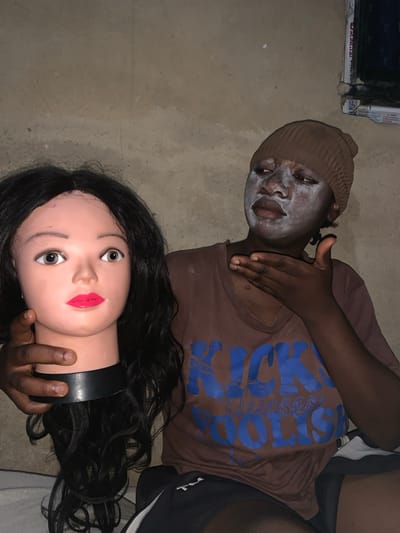 A medium shot depicts an individual and a mannequin head against a plain, possibly concrete, backdrop. The location is possibly Agban, Nigeria.
The individual, wearing a brown t-shirt with blue lettering, has a knit cap and a white substance applied to their face. Their right hand rests on their neck. The individual's expression is neutral.
A mannequin head with black hair is held by the individual's left hand. The mannequin has exaggerated makeup, including red lipstick.
The background is nondescript, appearing to be an interior space with what may be a window visible in the top left corner. The lighting is dim and even.