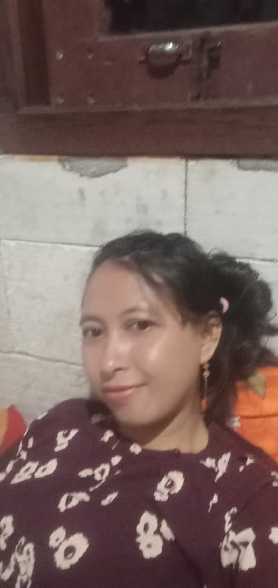 A medium close-up shows a woman indoors, possibly in Baron, Indonesia. She is the main subject, looking towards the camera with a slight, pleasant smile. Her dark hair is pulled back, with a few strands framing her face, and a pink hair tie is visible on the right side of her head. She wears dangle earrings and a long-sleeved shirt that is dark maroon or burgundy with light-colored, possibly white or cream, abstract or floral patterns.
The background consists of a light-colored wall with visible grout lines, resembling concrete blocks or large tiles. Some areas of the wall appear slightly worn or patched. Above the wall, a dark brown wooden structure, possibly a window or door frame, is visible, featuring a metallic latch or handle. On either side of the woman, parts of a red and orange pillow or fabric can be seen, adding a touch of color to the scene. The lighting suggests an indoor setting, possibly at night or in low light, with some sheen on her face from a light source. No visible text is present in the image.