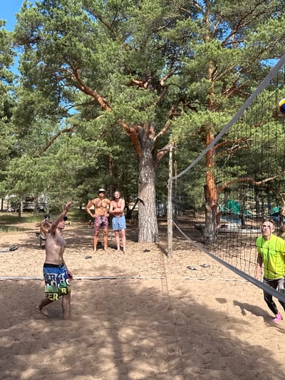 The image depicts a beach volleyball game taking place in Zelenaya Roshcha, Russia. Four adult males are visible on the sand court, with a volleyball net stretched across the frame. In the foreground, a man in patterned shorts is in mid-action, appearing to serve or hit the ball, with sand kicked up around his feet. To the right, partially obscured by the net, a man in a neon green shirt and patterned pants is looking towards the court. In the background, two men stand near the net; one in patterned shorts and a hat, and another in solid blue shorts. The environment is a sandy beach surrounded by tall pine trees, with what appear to be recreational vehicles and tents visible further in the background, suggesting a campground or recreational area. The sky is clear and blue, indicating a sunny day.