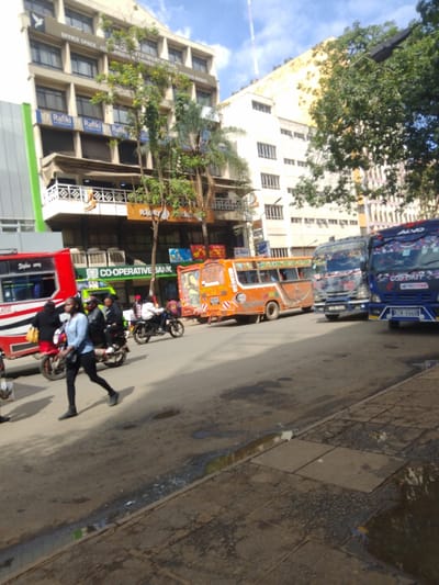 The image shows a street scene in Athi River, Kenya, located at latitude -1.453 and longitude 37.007. The street is paved with asphalt and has a sidewalk on the right. There are several vehicles parked on the street, including a red and white bus, an orange minibus, and a blue truck. There are also several people walking on the street and sidewalk, including a woman walking with a backpack. There is a building on the left side of the street, with a sign that says "CO-OPERATIVE BANK". There are several trees on the street and sidewalk, and the sky is clear. There is a sign with the words "Rafiki" repeated multiple times. The road is in rough condition with several large potholes. The image was taken during the day, and the lighting is bright.