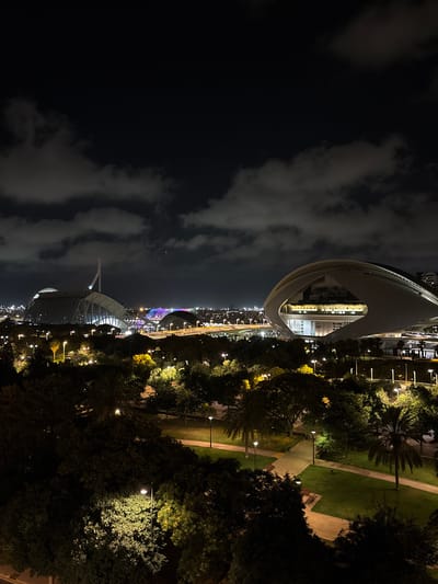 This image depicts a nighttime view of the City of Arts and Sciences in Valencia, Spain. The foreground is dominated by dark, leafy trees and parkland illuminated by scattered streetlights. In the midground, paved pathways and grassy areas are visible, with benches and lampposts providing light. The background features the distinctive architecture of the City of Arts and Sciences complex, including the Palau de les Arts Reina Sofía with its sail-like structure and the Hemisfèric with its eye-shaped design. Other illuminated structures and a colorful, arched bridge are also visible in the distance, along with a dark sky filled with scattered clouds. There are no people visible in the image.