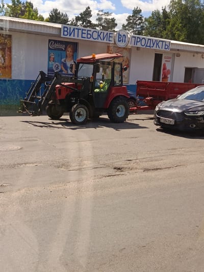 This image shows a red tractor with a front loader parked on a paved road in front of a building with a white corrugated metal facade. The building has blue text above it that reads "ВИТЕБСКИЕ ВО ПРОДУКТЫ" (Vitebskie VO Produkty), suggesting it is a food products store or company. On the building's wall, there are advertisements. One poster shows a young girl with pigtails holding a book, with the text "Сафійка" (Sofiyka) visible, likely a brand name. Another poster appears to advertise ice cream. To the right of the tractor, a red trailer is attached, and a dark-colored car with a license plate reading "1354 EM-2" is parked. The scene appears to be an outdoor setting, possibly a business or distribution center, in Vitsebsk, Belarus. The sky is partly cloudy, suggesting daytime. The tractor is positioned to possibly move or load goods, hinting at ongoing operations.