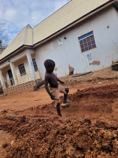 A medium shot captures a barefoot, dark-skinned child wearing light-colored shorts. The child stands beside a wall and a pile of red dirt. In the background is a two-story building with blue-framed windows. Scaffolding is visible in the background as well. The location appears to be Alizaga, Nigeria.