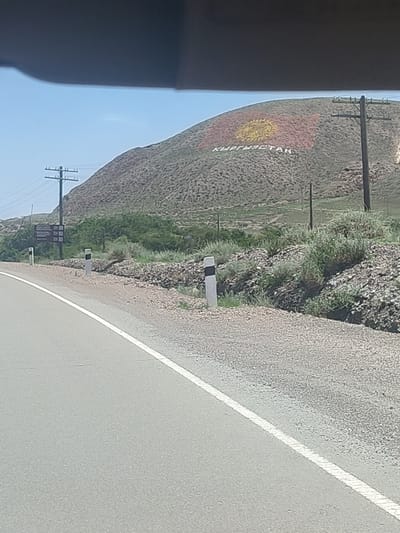 The image depicts a hillside in Kok-Moinok - 1, Kyrgyzstan, with a large representation of the Kyrgyz flag painted on its surface. Below the flag, the word "КЫРГЫЗСТАН" (Kyrgyzstan) is written in large white letters. The hillside is covered in dry vegetation and rocks. In the foreground, a paved road with a white line marking runs from left to right. Several road markers, black and white posts, are visible along the side of the road. Utility poles with wires are also present on the hillside and in the background. The sky is clear and blue.
