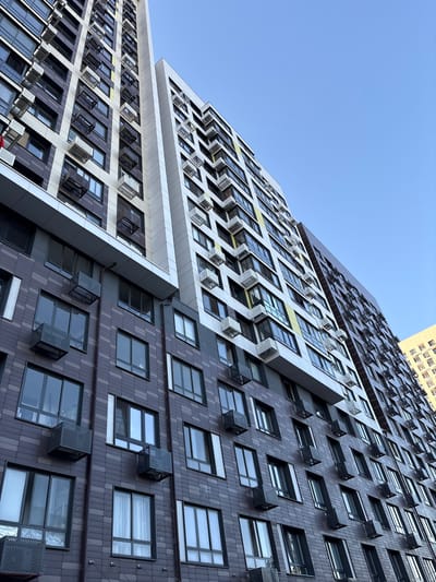 This media file features a low-angle, upward shot of a modern, multi-story residential building against a clear, blue sky, suggesting a daytime setting. The architecture is characterized by a staggered, asymmetrical design with contrasting facade materials, primarily dark gray tiles and white, angular panels. Numerous windows are visible, many equipped with external air conditioning units, some of which have protective grilles. There are no people visible in the image. The building appears to be located in a developed urban area, with another, older-looking building faintly visible in the upper right corner. The overall impression is one of contemporary urban architecture. The location context provided is Mytishchi, Russia. No text is visible in the image.