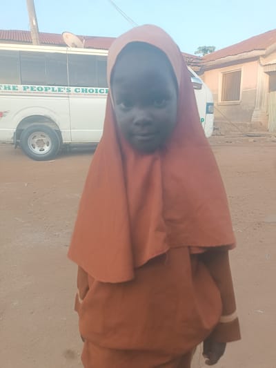 A young girl wearing an orange hijab and matching outfit stands in front of a white bus with "THE PEOPLE'S CHOICE" written on its side. The bus also has text indicating it is part of a "TARIA STATE MINISTRY OF CO OPERATION AND POVERTY ALLEVIATION PROGRAM." The girl's face is visible, looking towards the camera. The ground is dusty and sandy, consistent with an outdoor environment in Wukari, Nigeria. Buildings with corrugated iron roofs are visible in the background. The scene appears to be an everyday street view.