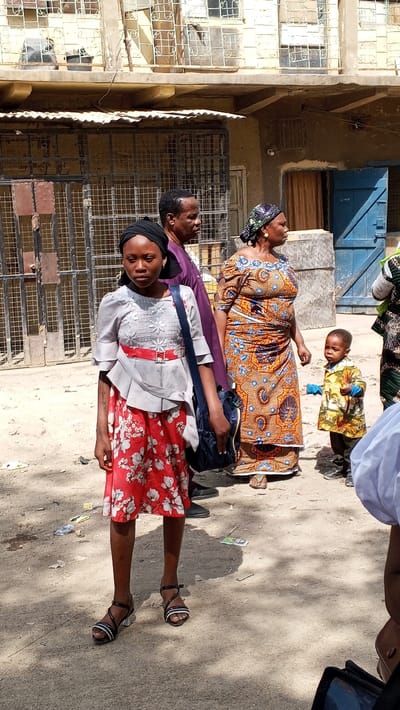 The image presents an outdoor scene in Sabon Gari East, Nigeria, under daylight conditions. The ground surface is primarily light brown dirt with scattered small pieces of debris. Five individuals are visible, four of whom are fully or largely framed.

In the foreground, slightly left of center, a young female stands, facing slightly right with a neutral expression. She wears a dark head covering, a light gray short-sleeved top featuring a red sash with a buckle and white embroidery, and a red skirt with a white floral pattern. A dark blue shoulder bag is worn across her body, and she wears black and white sandals.

Behind her, a male individual stands in profile, facing right, dressed in a dark purple garment. Adjacent to him on the right, a female individual stands in profile, also facing right. She wears a patterned headwrap and a long, patterned dress in shades of orange and blue, with sandals visible on her feet.

To the right of the second female, a young child stands, facing slightly left. The child wears a yellow patterned short-sleeved shirt, dark trousers, and dark shoes, holding a small, indistinct object in its left hand. The right shoulder and arm of a fifth individual, wearing a white garment, are partially visible in the bottom right corner of the frame.

The background consists of a two-story building with a light-colored facade. The ground floor features a large metal gate on the left and a blue wooden door on the right, with a corrugated metal awning extending above. The upper floor displays multiple windows secured by decorative metal grilles. Miscellaneous objects are visible on the ledge or balcony of the upper floor. Shadows are cast on the ground, consistent with direct sunlight.