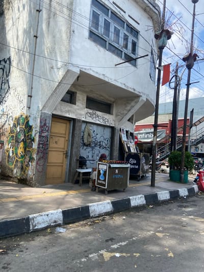 The image shows a street corner in Cimahi, Indonesia (latitude: -6.874, longitude: 107.542). A man is sitting on a small stool in front of a wooden cart selling merchandise. The cart has a sign that says "Efis Agura" and "N Special Santika." The man is wearing a long-sleeved shirt and a hat. He is looking down at the cart. The cart is in front of a white building with a large window and a door. The building has graffiti on the exterior. There is a red sign with the word "Infinix" on it on the building. The street is paved with asphalt and there are white lines painted on the side of the road. There are two tall poles on the right side of the image, with decorations on top of them.  There is a red flag hanging from one of the poles. There is also a small bush and a motorbike parked on the side of the street. The background is a busy street with other buildings and cars.