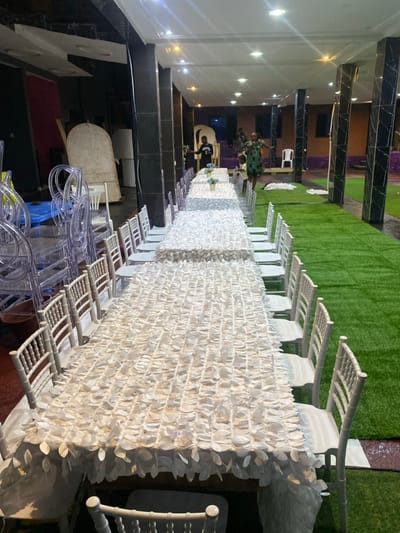 The image shows a long table covered in a white tablecloth with a decorative ruffled design, set with white chairs on either side. The table is situated in an outdoor setting, likely a garden or patio, featuring artificial turf and a few potted plants. The location, based on the coordinates, is in the city of Limbe, Cameroon. There are multiple people visible in the background, seemingly preparing for an event. The scene is well-lit, highlighting the table and chairs, creating a festive ambiance. The image focuses on the table setup, suggesting a gathering or celebration.