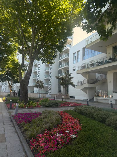 The image depicts a modern hotel complex in Obrochishte, Bulgaria. The foreground features a well-maintained garden with vibrant flowers in shades of pink, red, and purple, interspersed with lush green foliage. A paved walkway runs alongside the garden. Behind the garden, a large tree with broad leaves provides shade. In the background, a multi-story white building with contemporary architecture dominates the scene. The building has multiple balconies with glass railings and outdoor seating areas. A prominent external staircase leads up to one of the building's levels. The sky is a clear blue, suggesting a sunny day. There are no people visible in the immediate foreground or on the balconies. The overall impression is one of a peaceful and well-kept resort.