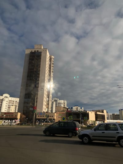 The image depicts a street scene in Mahilyow, Belarus, on a cloudy day with the sun glinting off a tall building. The perspective is from a low angle, looking upwards towards the sky and buildings.
Dominant subjects include several multi-story buildings, some with modern glass facades and others with more traditional architecture. A light-colored SUV and a dark sedan are parked or driving on the paved street. The sky is filled with a mix of dark and light clouds, suggesting overcast conditions with intermittent sunshine.
The scene appears to be a commercial or residential area, with various signs visible on the buildings. Some of the text on the signs is in Cyrillic script. One prominent sign reads "ГЛАВЕВ МОЙ ГОРОД" which translates to "GLAVEV MY CITY". Another sign with an "АПТЕКА" (Pharmacy) logo is also visible. The presence of streetlights and crosswalk markings indicates an urban environment.
The time of day is difficult to determine precisely due to the cloud cover, but the sun's reflection suggests it is daytime, likely afternoon. There are no people clearly visible in the image. The overall mood is neutral, focusing on the architecture and the urban landscape under a dynamic sky.