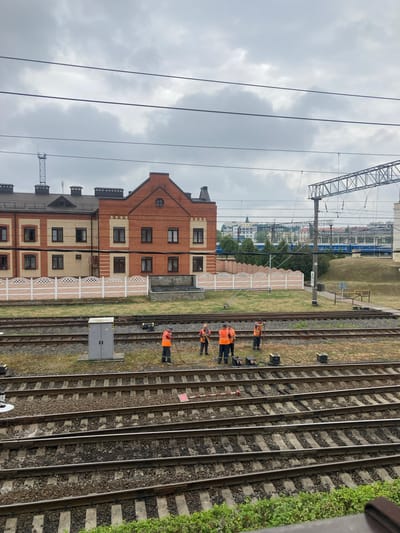 The image depicts a cloudy day at a train station in Brest, Belarus. In the foreground, several workers in orange high-visibility vests are engaged in maintenance work on the train tracks. They are using shovels and other tools, suggesting they are clearing debris or performing repairs. There are several electrical boxes and equipment present near the tracks. In the middle ground, a modern building complex with brick and beige facades is visible behind a white decorative fence. Further in the background, a blue train is visible on the tracks, and beyond that, more buildings and trees form part of the urban landscape. Overhead, electrical wires for the trains crisscross the sky.