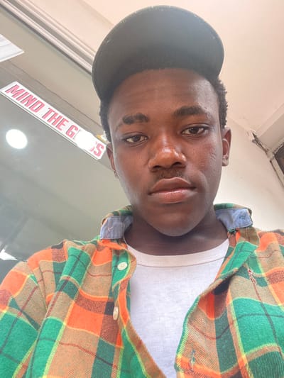 The image displays a close-up, chest-up portrait of a young male individual, presumably taken in Ikeja, Nigeria. The subject has dark skin, short dark hair, and wears a dark baseball cap. He looks directly forward into the camera. A faint mustache is visible above his upper lip. He is dressed in an unbuttoned plaid shirt featuring orange, green, and dark brown/black patterns, worn over a plain white t-shirt with a light blue collar visible. The background is out of focus but shows a light-colored ceiling overhead. To the left, a reflective surface, likely glass, features a red sign with white text partially visible, reading "MIND THE G**S," suggesting "MIND THE GLASS." A circular light source is reflected in this surface. To the right, a light-colored wall or column shows signs of wear, including peeling paint or plaster. No other individuals are present, and the subject appears to be taking a selfie or being photographed at close range.