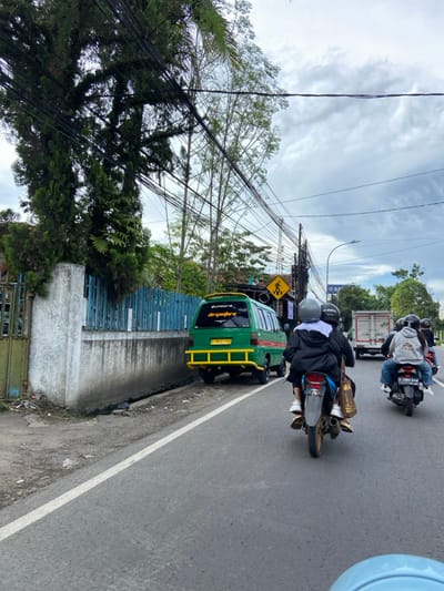 The image shows a street in Katapang, Indonesia (latitude: -7.007, longitude: 107.551). A green van is parked on the right side of the road, facing the opposite direction. Two motorbikes are riding on the road. The rider of the first motorbike is wearing a black jacket and helmet, and the rider of the second motorbike is wearing a blue jacket and helmet. Both motorbikes are heading in the same direction. The street is bordered by a white wall and a row of trees on the left side. The road is paved and there are numerous power lines overhead. The weather appears overcast. There are no pedestrians visible.