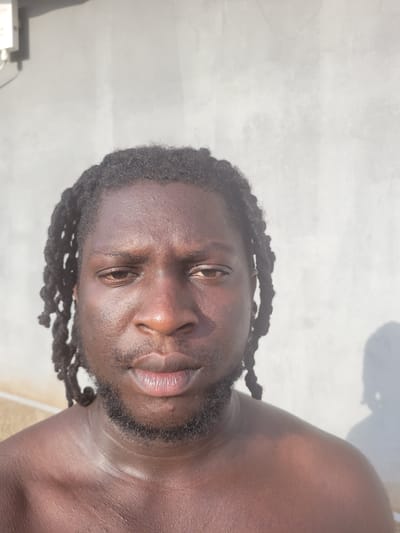 A close-up, angled portrait of a man with dark skin, dreadlocks, and a beard. He appears to be shirtless and is likely outdoors. The background is a light gray, possibly a concrete surface or wall. No specific details indicate the location as Markurdi, Nigeria.