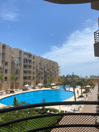 View from a high vantage point, potentially a balcony, showing a multi-story hotel building next to a swimming pool in Hurghada, Egypt. The building is beige, with multiple rows of windows and balconies. A large, oval-shaped blue swimming pool dominates the foreground. People are visible, either swimming or sunbathing around the pool. Palm trees are interspersed between the pool and the building. Clear blue sky is visible above the building. A section of a metal fence is visible on the right side of the image.