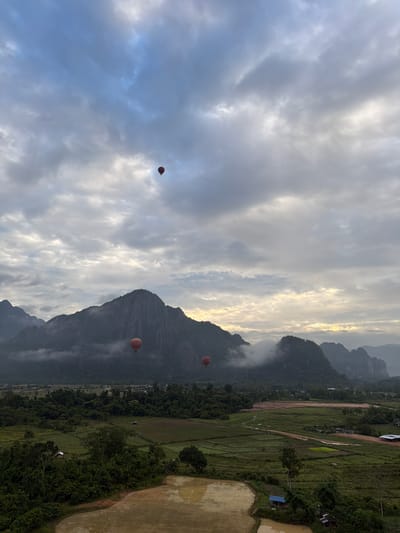 In Vang Vieng, Laos, a scenic landscape unfolds under a cloudy sky, with a majestic mountain range forming the backdrop. The mountains, partially shrouded in mist, rise dramatically from the lush green terrain below. In the foreground, cultivated rice paddies reflect the muted light of the overcast sky. Scattered throughout the landscape are dense patches of trees and a few small structures, hinting at a rural settlement. The scene is further enlivened by the presence of several hot air balloons floating gracefully in the sky. One balloon, a vibrant red, is prominently positioned in the upper left portion of the image, while two others are smaller and further away, one near the mountains and another higher in the sky. The overall impression is one of a serene morning in a picturesque location.