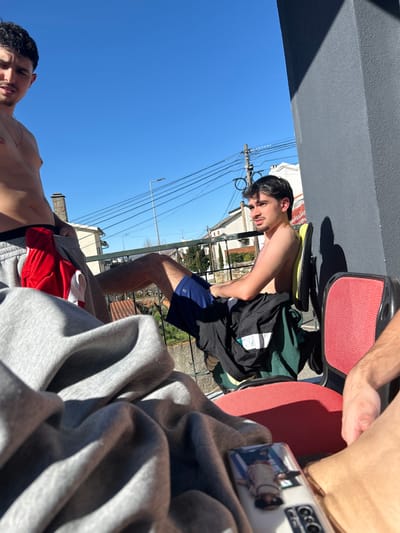The image captures three individuals outdoors on a sunny day within a residential area of Bragança, Portugal.
In the left midground, a young male with dark hair and a mustache stands shirtless, wearing grey athletic shorts with a red garment partially visible inside. He faces the camera. A faint mark or scar is visible on his left chest.
To the right, a second young male with dark hair and a mustache is seated shirtless in a black office chair featuring a yellow-green backrest. He wears dark blue athletic shorts, and a black t-shirt with a white stripe is draped over the chair. He gazes towards the viewer's left. His left leg is extended forward.
In the immediate foreground, a substantial amount of grey fabric, consistent with clothing or a blanket, is positioned on the left. On the right, a white smartphone, encased in clear plastic displaying an inverted image of a person, rests on a red surface, likely the seat of a third chair. A partial view of a third individual's arm and leg is visible to the right of the smartphone.
The background features a clear blue sky. A metal railing separates the subjects from a residential panorama of houses with tiled roofs, one featuring a prominent stone chimney. Overhead utility lines, a street lamp, and sparse deciduous trees are present. A dark grey vertical structure on the far right casts a sharp shadow, indicating direct sunlight. A distant building displays a partial sign reading "eissell" or "elssele" followed by "serm" or "term". The setting appears to be a balcony or terrace.