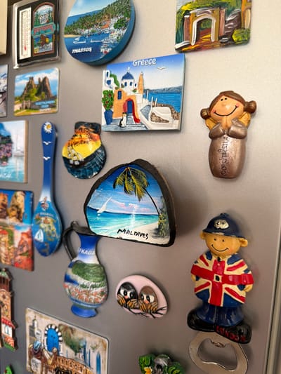 The image displays a collection of magnets on a metallic surface, likely a refrigerator door. The magnets feature various tourist attractions and souvenirs from different locations. Notable magnets include a thermometer magnet for "West Virginia," a round magnet depicting "THASSOS," a rectangular magnet with a Greek village scene labeled "Greece," and a shell-shaped magnet with a ship and the text "SAINT HELENA." Other magnets show a palm tree beach scene with "MALDIVES" written on it, a blue spoon magnet with various illustrations, a jug magnet with a landscape and the text "FKADA," a small oval magnet with two owls, and a magnet depicting a camel and a blue doorway. There is also a figurine of a person in a police uniform wearing a Union Jack-themed coat, with "LONDON" written on their shoes, which also functions as a bottle opener. Another magnet is a figurine of a person with their hands clasped in prayer. The overall environment is cluttered with these decorative magnets. The city of Pazardzhik, Bulgaria is not depicted in this image.