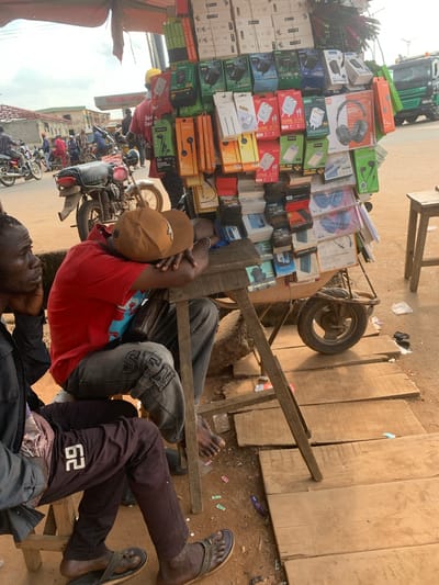 This media file captures a busy street scene in Shagamu, Nigeria, likely during the daytime, given the bright lighting. The foreground is dominated by a man wearing a red t-shirt, a beige baseball cap, and jeans, resting on a makeshift wooden structure. He appears relaxed, with his arm resting on his head and a brown bag beside him. Another person, wearing dark pants with the number "62" prominently displayed, is also seated.
Behind the seated individuals, a market stall is packed with an assortment of electronics, primarily headphones and chargers in colorful packaging. Prominent text includes brand names like "JBL," "AREGOLD," and "Total Energies." The stall is built on a cart with a large wheel, suggesting it might be mobile.
In the background, a street is filled with various motorcycles and a few other individuals. The general atmosphere suggests a bustling commercial area. The presence of a large truck on the left side of the image indicates ongoing traffic. The scene conveys a sense of everyday life and commerce in a Nigerian urban environment.